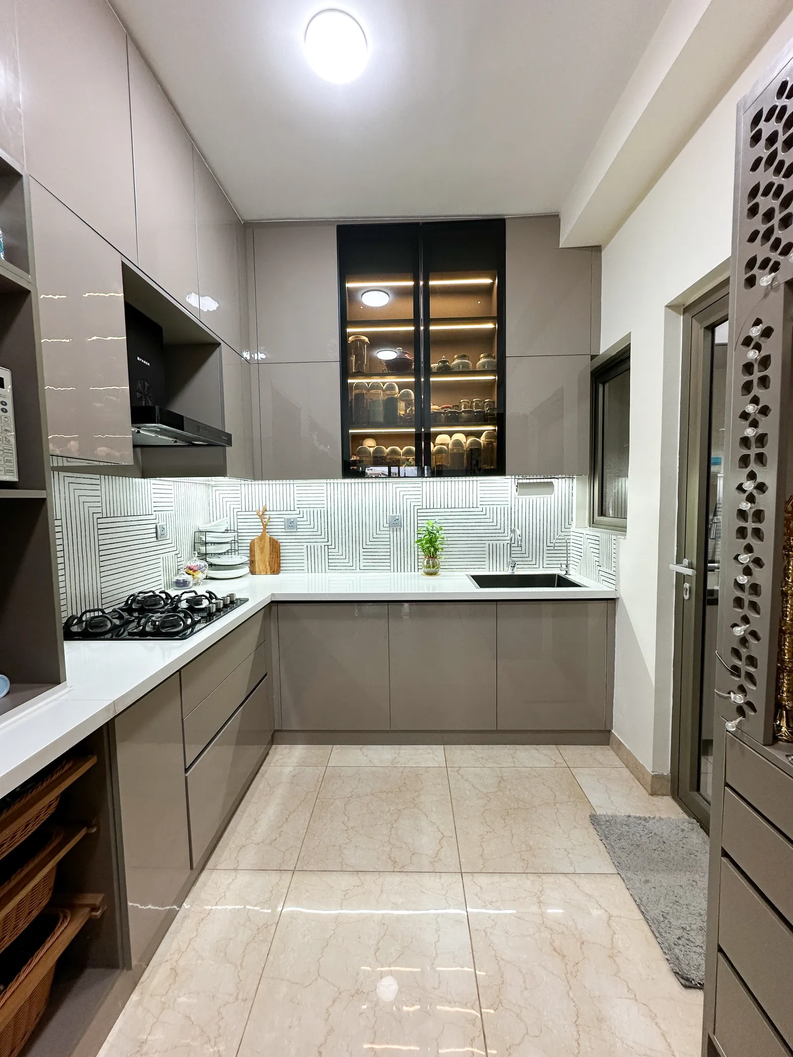 L-shaped kitchen in taupe with lit glass-front tall unit — Somani's Residence